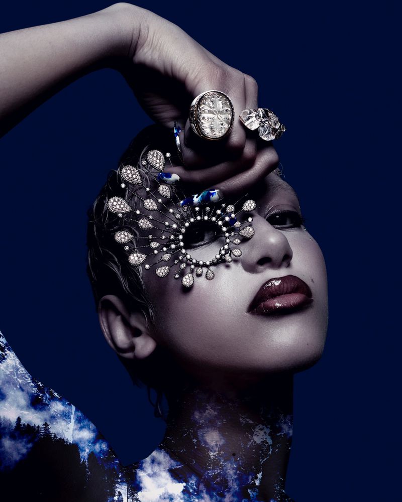 Glamorous woman in artistic makeup and jewelry poses against dark backdrop in a beauty or fashion concept image with blue tones.