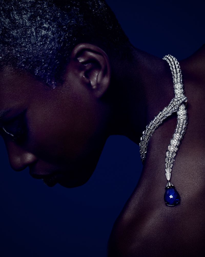 Close-up fashion shot of a woman with an elegant diamond necklace featuring a large blue gemstone pendant against a dark blue backdrop.