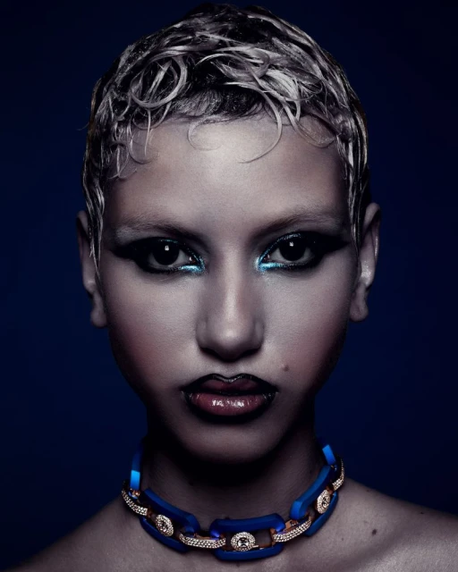 Close-up portrait of a woman with short, wet-look hair, dramatic eye makeup, dark lipstick, and a blue chain necklace against a dark background.