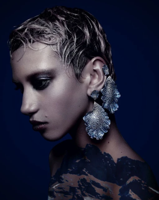 Eye-level shot of a fashion model with short hair wearing large leaf-shaped statement earrings in front of a solid dark blue backdrop.
