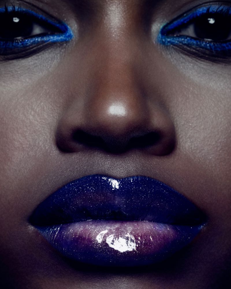 Close-up of a woman's face with dark skin, wearing blue eyeshadow and dark purple lipstick with a glossy finish in soft lighting.