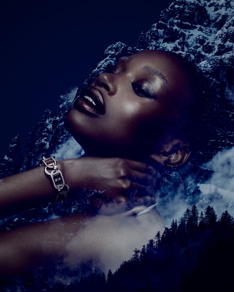 A woman with brown skin poses with a bracelet against a background with mountains and trees in a low angle, close-up studio shot.