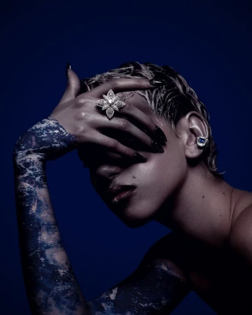 Model with wet hair wearing a diamond star ring and sapphire ear cuff against a dark blue background for Nu Icons.