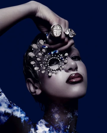 Close-up portrait of a woman wearing an intricate diamond eye mask, rings, and blue patterned body paint against a dark background
