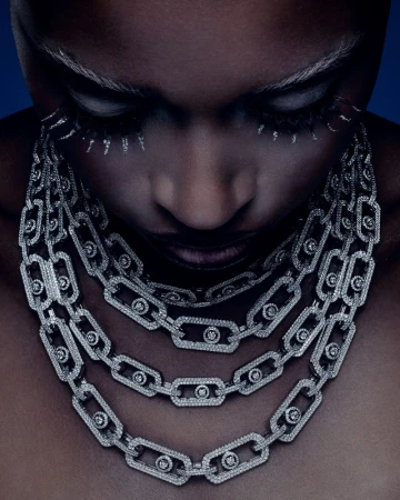 Close-up portrait of a woman wearing layered diamond chain necklaces with dramatic white eyelashes against a dark blue background.