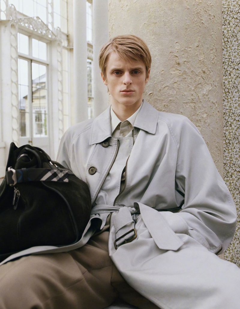 A blond male model in a trench coat poses against a marble column, holding a black duffel bag in an architectural setting.