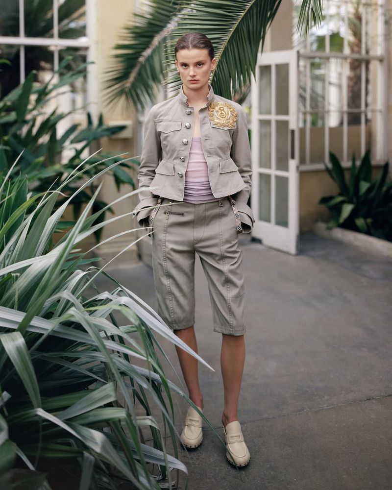 A full shot of a female model in a khaki shorts suit, posing in a garden setting with tropical plants and a building visible behind her.