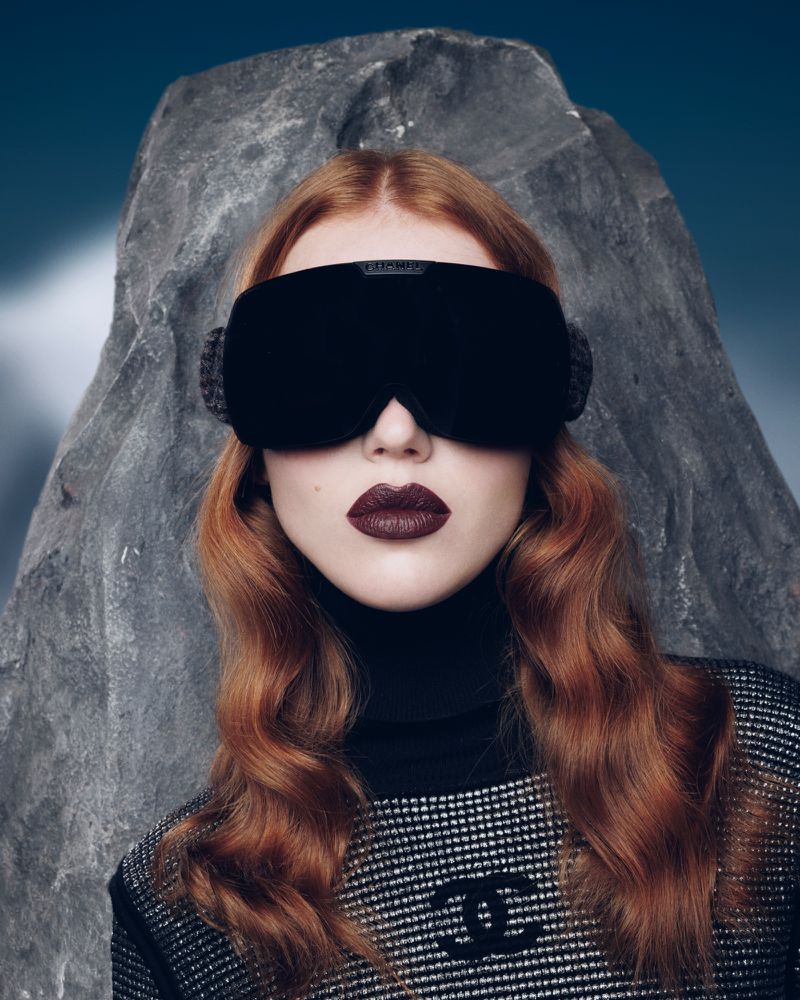 A red-haired woman wearing dark goggles and lipstick is the main focus, with a grey rock formation and blue backdrop adding depth to the shot.