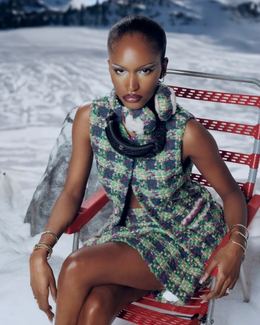Woman poses in patterned skirt suit on red chair in snowy mountain landscape. She wears ski goggles around her neck. Fashion shot, outdoor setting.