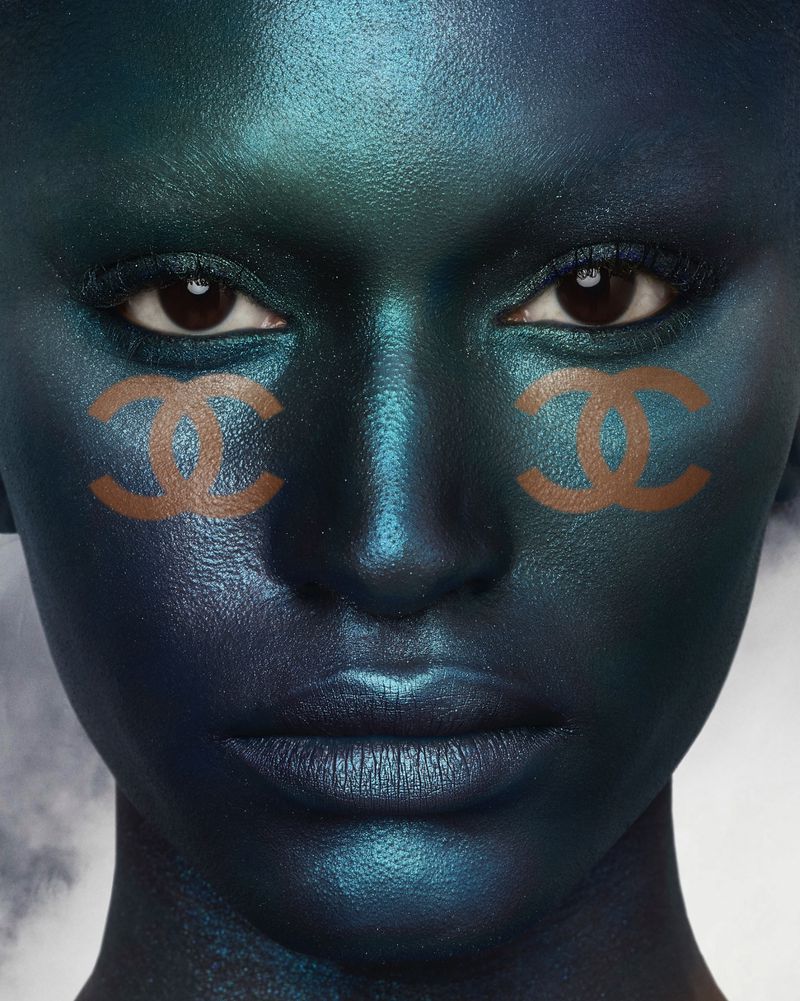A close-up studio portrait of a person with dark skin painted in blue and green tones, with Chanel logo face-paint near the eyes.