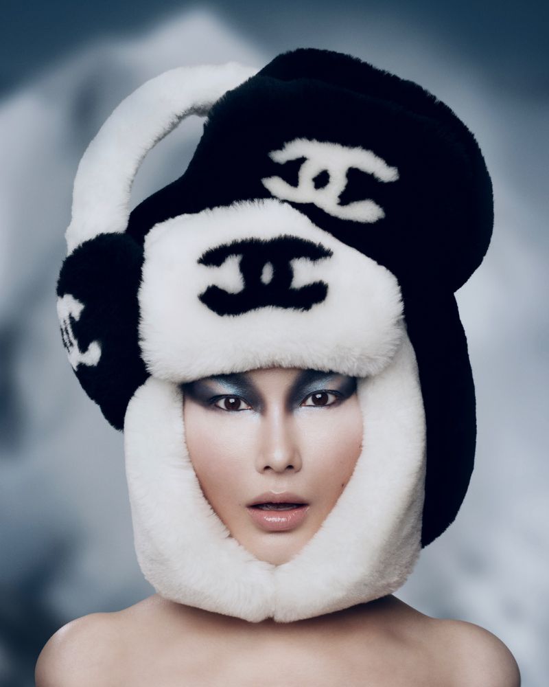Close-up portrait of a woman with theatrical silver makeup wearing a black and white fur hat with brand logo against blurred snow background.