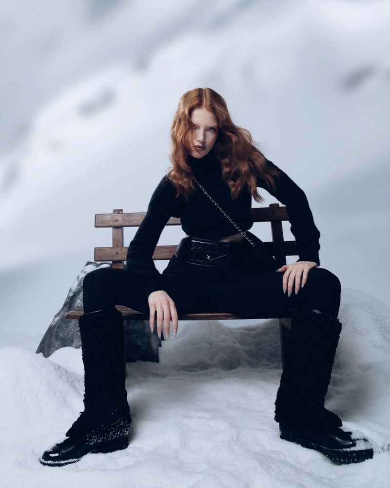 A woman with red hair sits on a wooden bench in the snow, wearing a black sweater, pants, boots and purse with a snow-covered background.