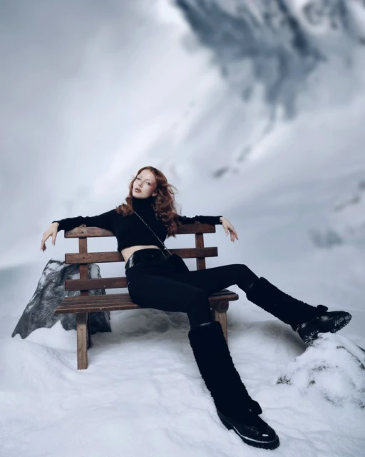 A woman sits on a wood bench wearing a black crop top, pants and boots with snow boots in a snow covered mountain landscape.