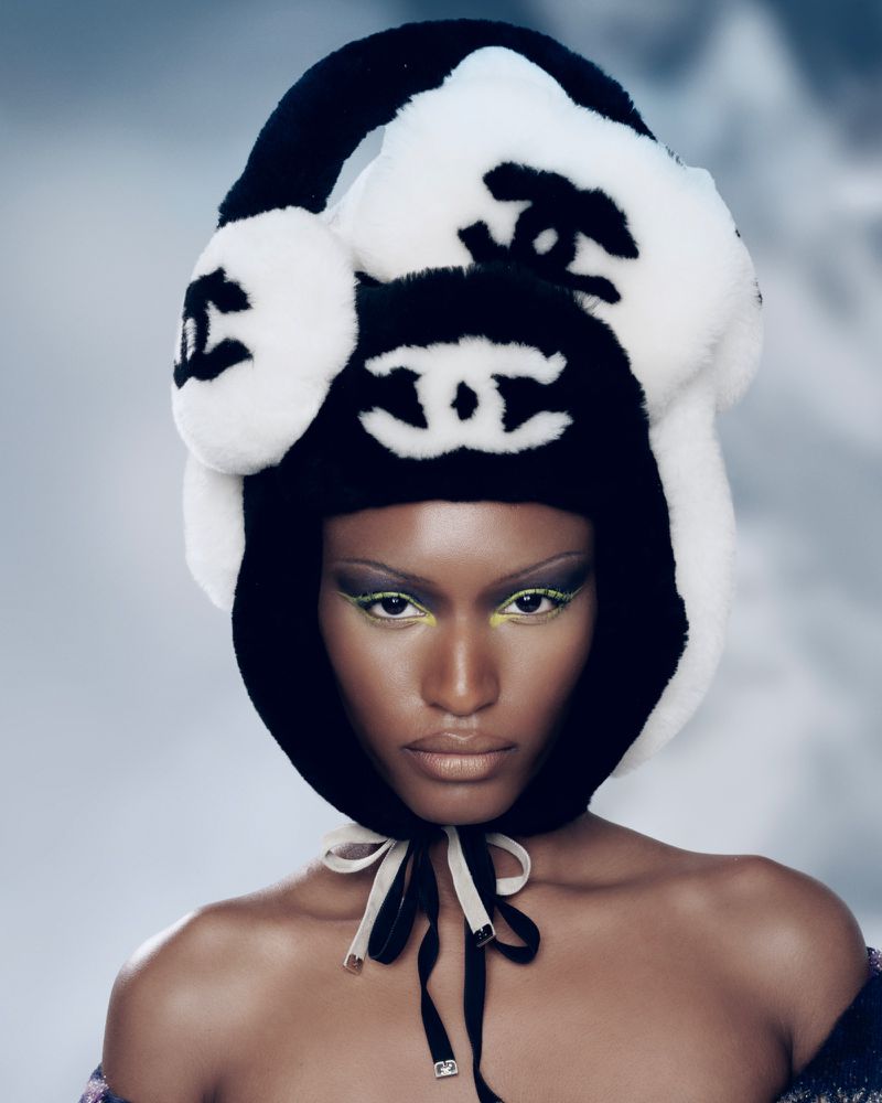 Close-up of a woman with dark skin wearing a black and white logo winter hat with green eyeshadow in a beauty fashion shot.