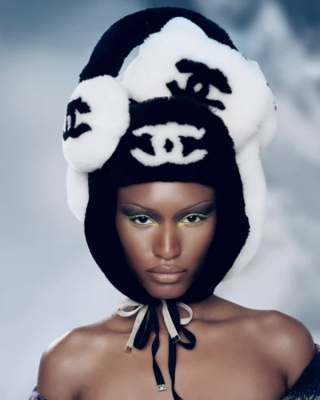 Close-up of a woman with dark skin wearing a black and white logo winter hat with green eyeshadow in a beauty fashion shot.