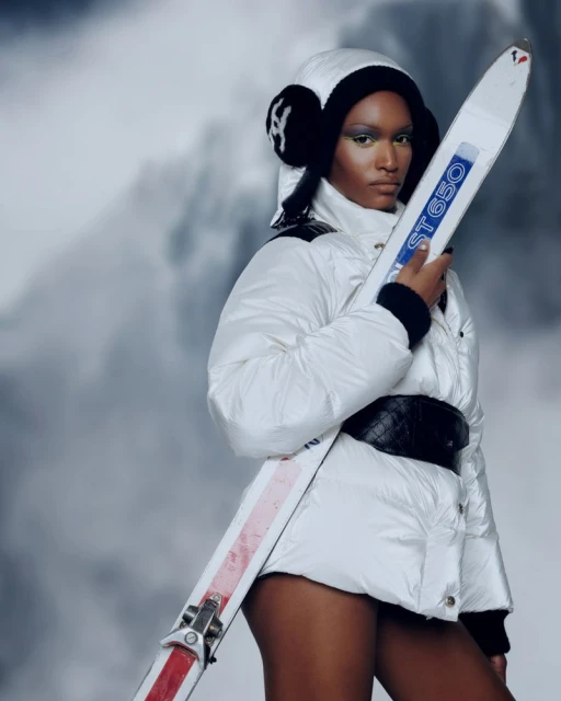 Woman in white ski suit and earmuffs holds skis against a blurred mountain background, showcasing winter fashion and athletic style.
