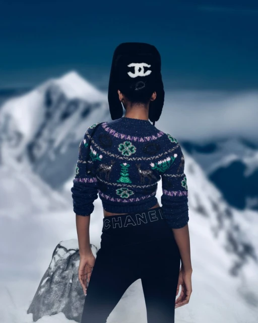 Back view of a person in a cropped knit sweater and fur hat stands on a snowy peak with a blurred snow-covered mountain range background.