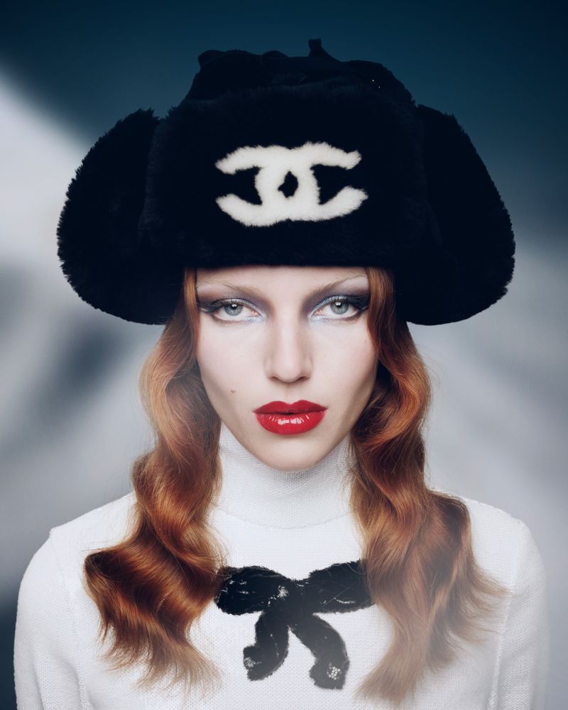 Fashion model wearing a fur hat with the Chanel logo, red lipstick, and a white sweater with a black bow against a blurry background.