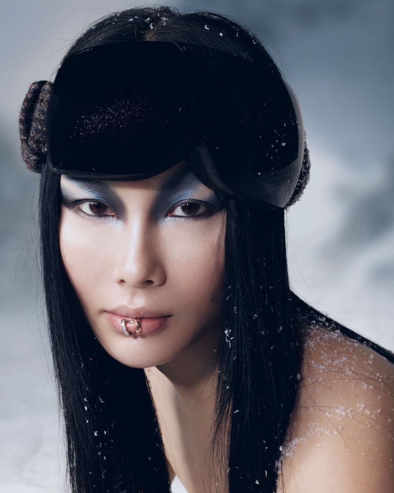 Portrait of an Asian woman with lip piercings wearing black ski goggles with a snowy background, dramatic eye makeup, and sleek hair.