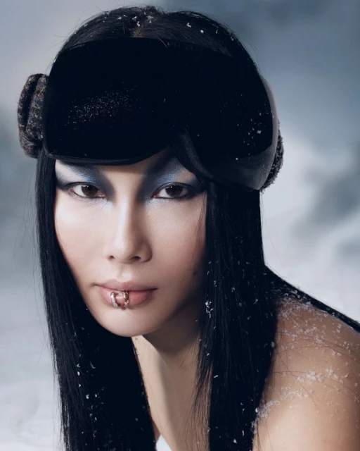 Portrait of an Asian woman with lip piercings wearing black ski goggles with a snowy background, dramatic eye makeup, and sleek hair.