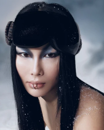 Portrait of an Asian woman with lip piercings wearing black ski goggles with a snowy background, dramatic eye makeup, and sleek hair.