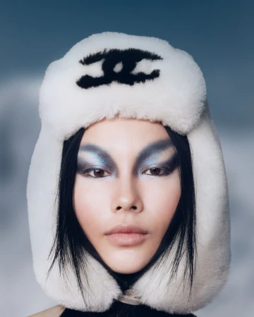 Headshot of a model wearing a white furry hat with a black Chanel logo, styled with dramatic blue eyeshadow, against a soft blue background.