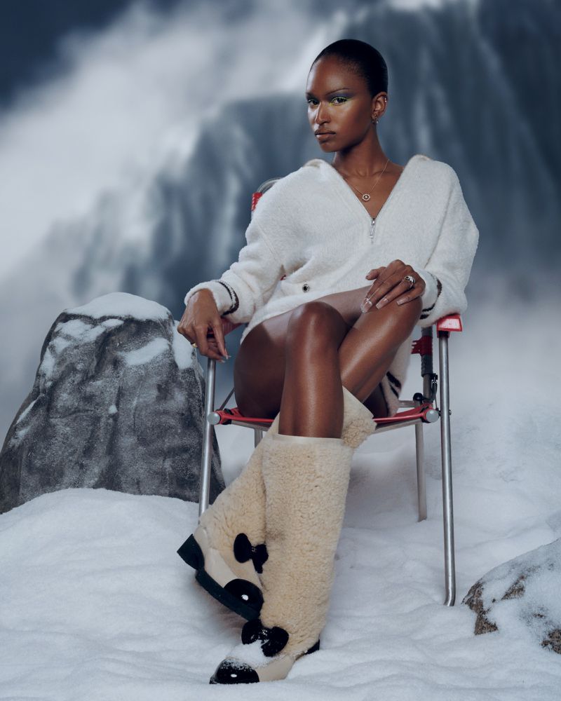 A woman with gold eyeshadow and fuzzy boots sits on a red folding chair against a scenic mountain backdrop in a studio setting.