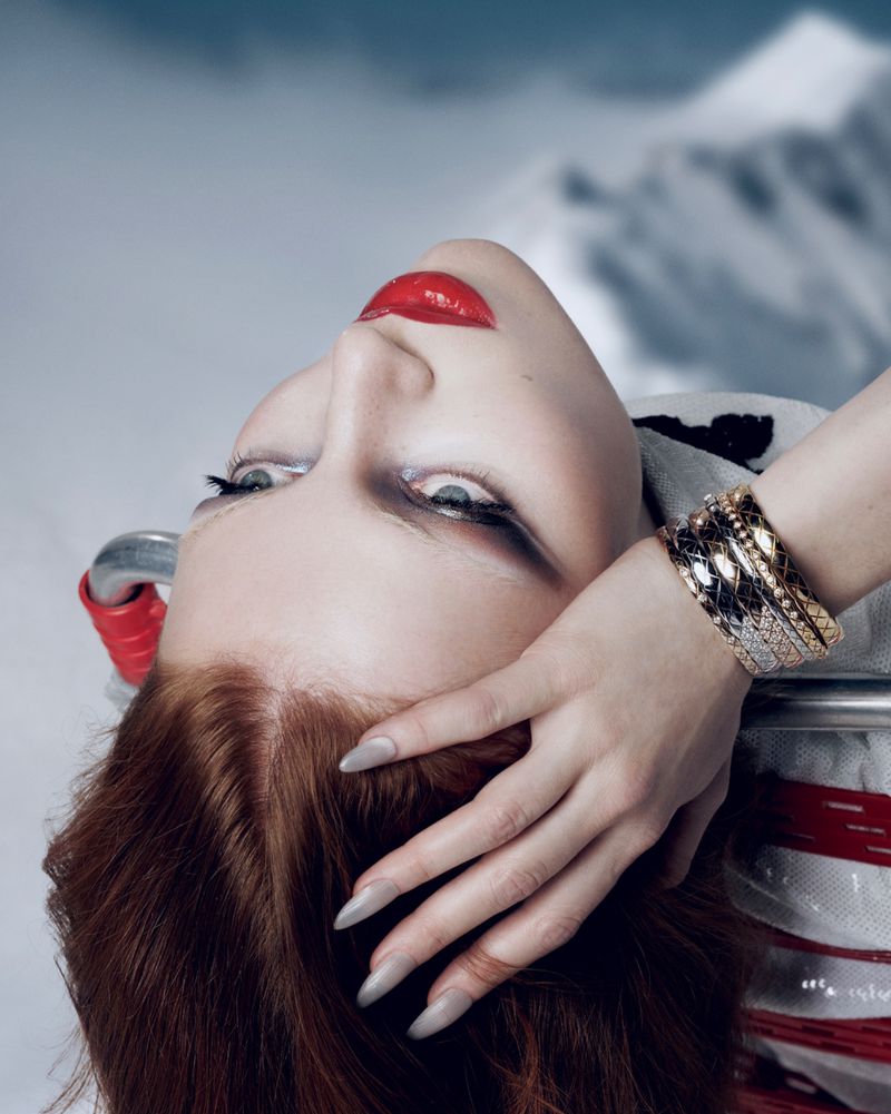 Fashion shot of a red-haired woman with red lipstick wearing gold bracelets in front of a blurred mountain background.