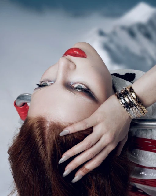 Fashion shot of a red-haired woman with red lipstick wearing gold bracelets in front of a blurred mountain background.