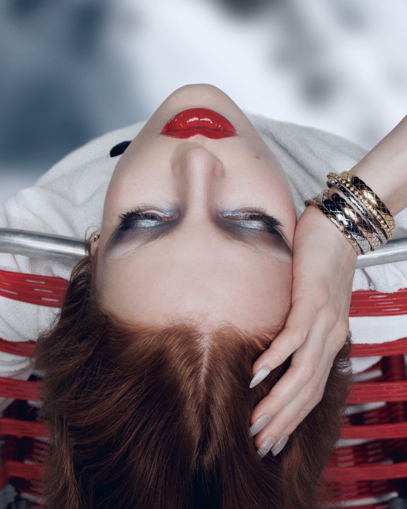 Glamorous portrait of a red-haired woman with striking silver eyeshadow and bold red lips, reclining with her hand on her head.