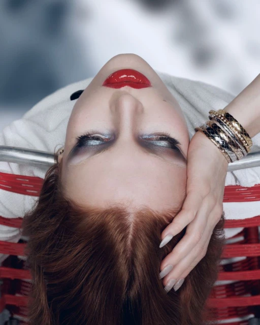 Glamorous portrait of a red-haired woman with striking silver eyeshadow and bold red lips, reclining with her hand on her head.
