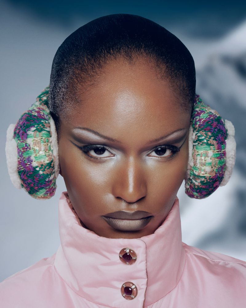 Close-up portrait of a Black model with high-fashion makeup, winter earmuffs, and a pink puffer jacket against a snowy mountain backdrop.