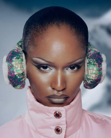 Close-up portrait of a Black model with high-fashion makeup, winter earmuffs, and a pink puffer jacket against a snowy mountain backdrop.
