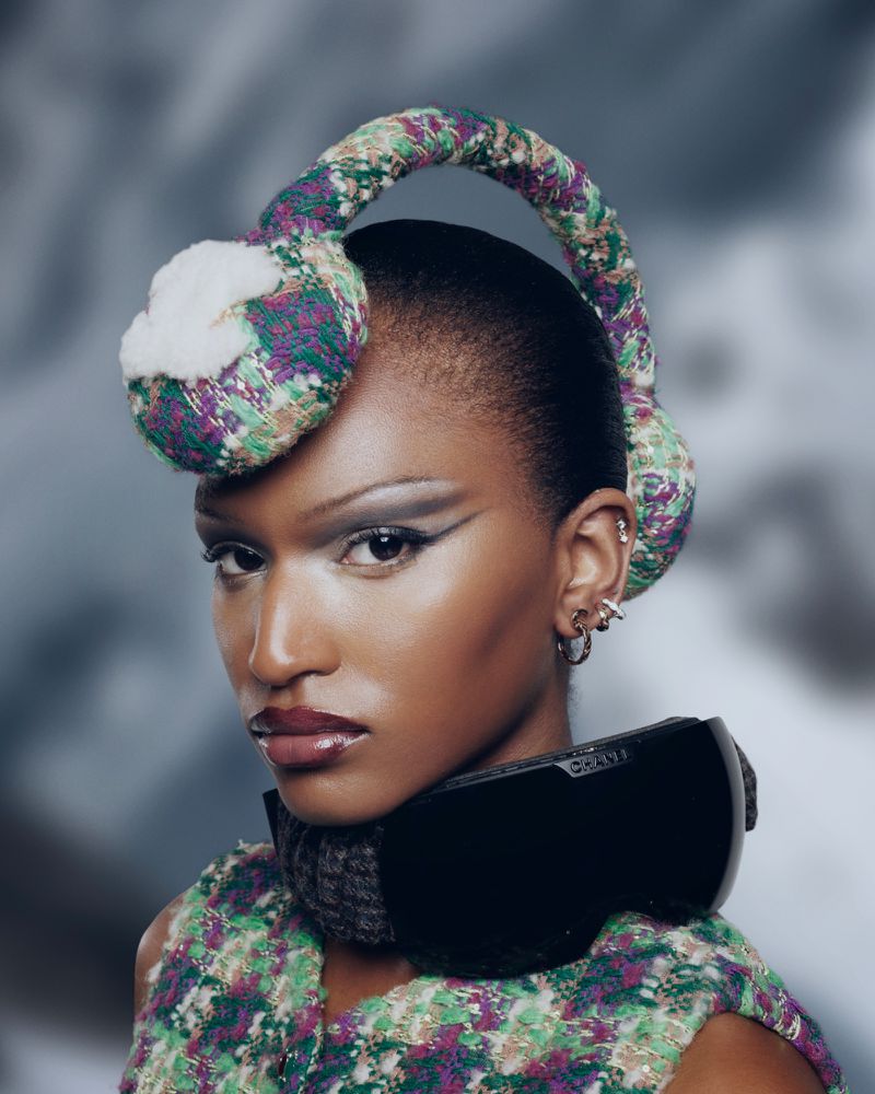 Portrait of a Black woman with stylish earmuffs and ski goggles, dramatic eye makeup, and glossy lips against a blurred winter backdrop.