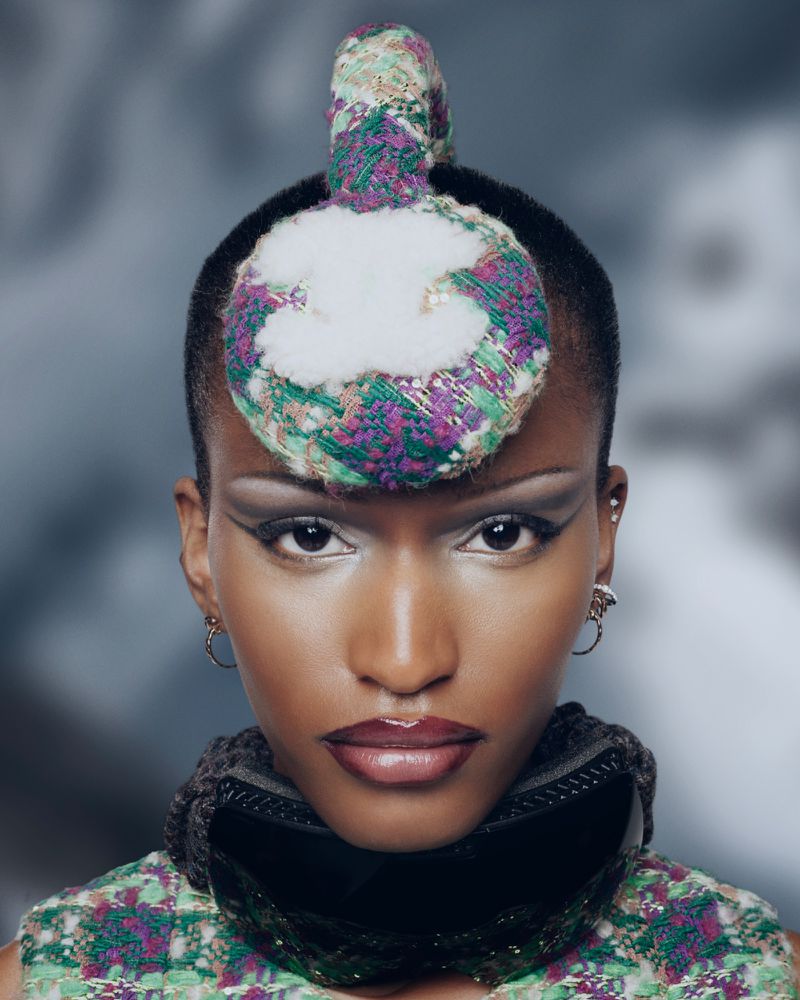 Close-up portrait of a black female with a unique, colorful headpiece, wearing a patterned top and a dark neck accessory against a blurred background.