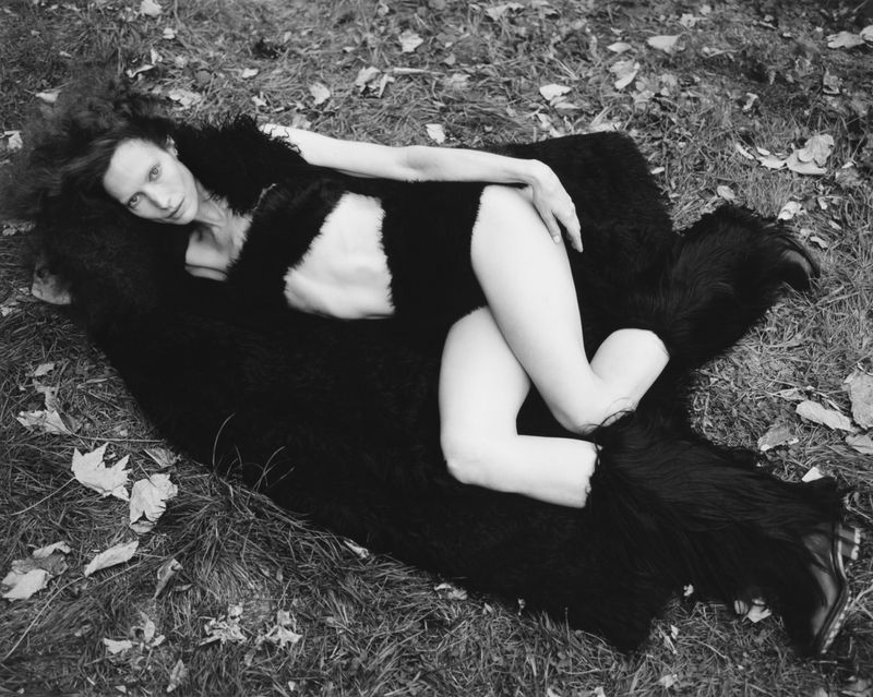 Monochrome shot of a slender woman in lingerie, lying on a fur rug in a grassy outdoor setting, creating a sensual, earthy fashion image.