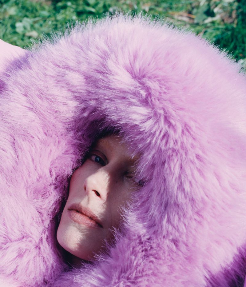 Portrait of a fair-skinned woman with blue eyes, partially framed by a large pink faux fur hood in a soft, natural outdoor setting.