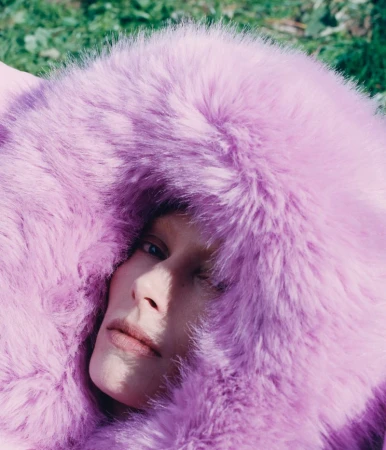 Portrait of a fair-skinned woman with blue eyes, partially framed by a large pink faux fur hood in a soft, natural outdoor setting.