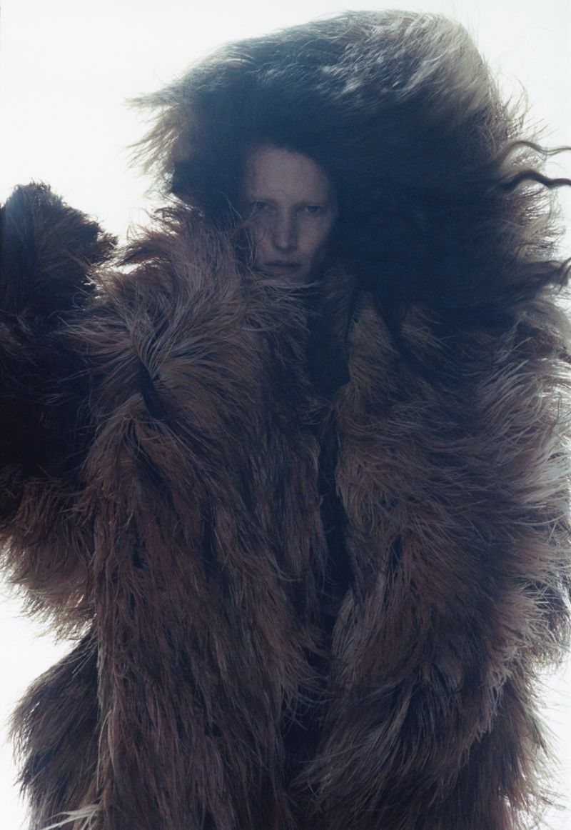 Person in a large, shaggy fur coat. The person has fair skin and is looking at the camera. White background. Studio lighting.