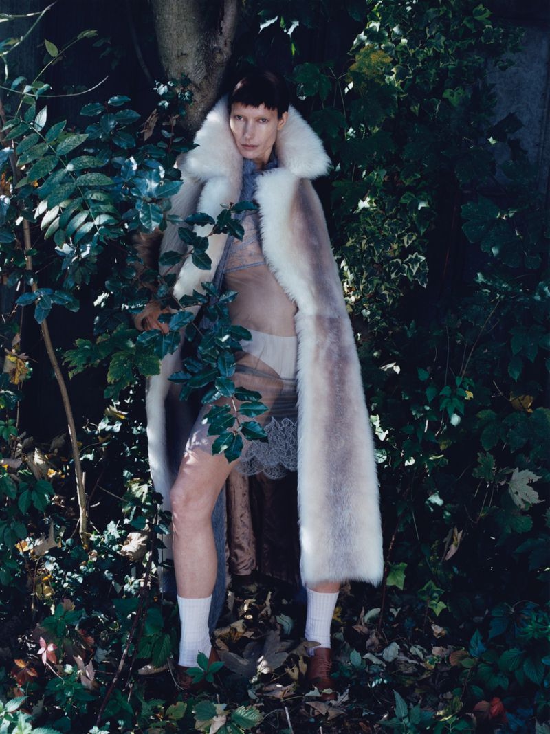 Woman poses in an outdoor setting wearing a long fur coat, sheer dress, brown shoes, and white socks surrounded by lush greenery.