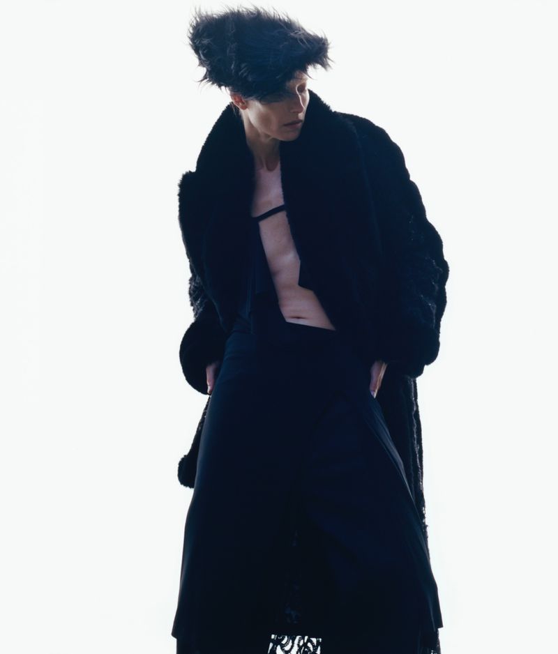 Person in oversized fur coat and hat stands against a white background in a fashion shot with hands in pockets, slightly looking away.