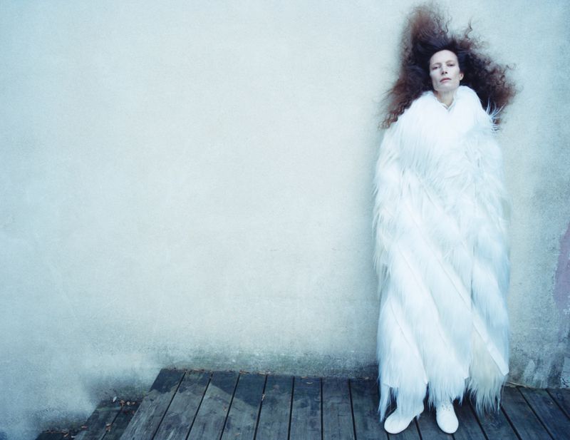 Adult female stands on wooden planks, covered in a long white faux fur coat against a mottled white wall. Auburn hair is spread out.