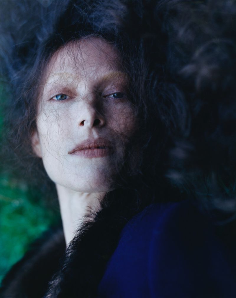 A close-up portrait captures a female with pale skin, blue eyes, and dark hair against a blend of dark blue and green backdrop.