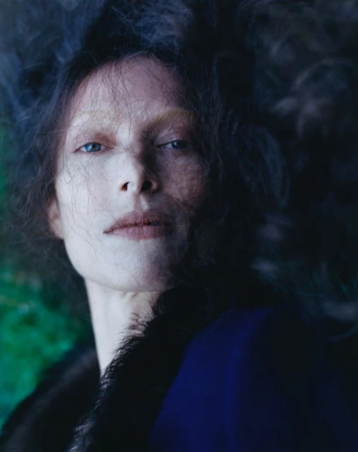 A close-up portrait captures a female with pale skin, blue eyes, and dark hair against a blend of dark blue and green backdrop. 