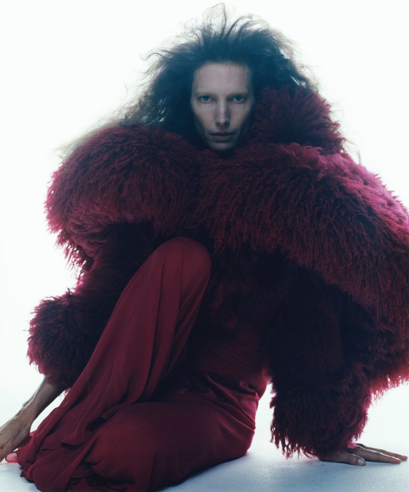 Fashion portrait of a person in a burgundy dress and a large fur-like coat, posing indoors against a bright, neutral background.