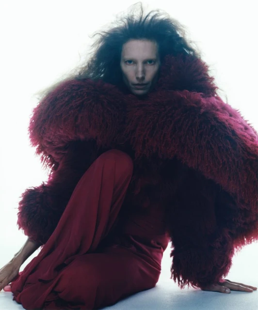Fashion portrait of a person in a burgundy dress and a large fur-like coat, posing indoors against a bright, neutral background.