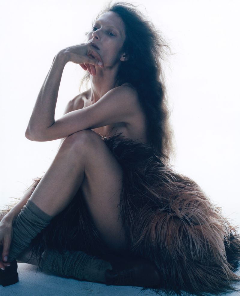 Fashion photo of a slim adult female model with long dark hair, sitting in a draped brown fur garment against a bright white backdrop.