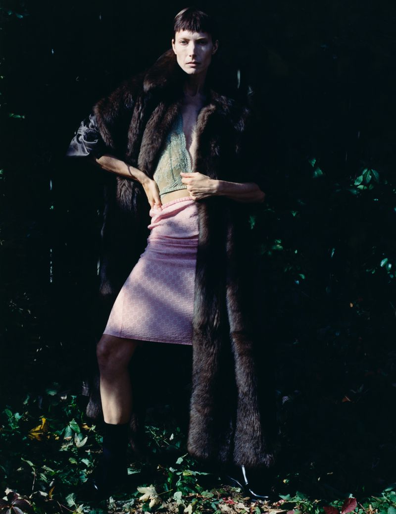 Model standing outdoors wearing a long dark fur coat, pink skirt, and knitted top against foliage