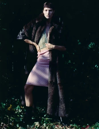 Model standing outdoors wearing a long dark fur coat, pink skirt, and knitted top against foliage
