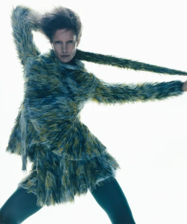 A fashion model poses dynamically in a textured blue and yellow faux fur jacket against a stark, high-key white background.